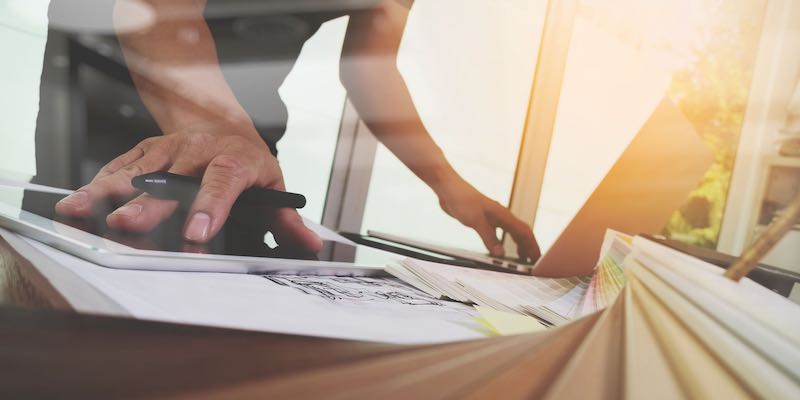 Mile One: Hard Work. Close up image of a man, pen in hand, working at a computer with a tablet and papers under one hand. The sun is causing a lens flare in the upper-left hand corner of the image, which adds to the warm tone of the photo.