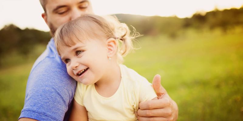 Mile Three: Integrity. Photo of a small child in her father's arms, outdoors in late afternoon, in a grassy, meadow.